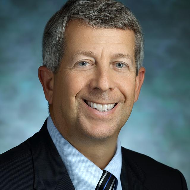 Andrew Satin, in a formal portrait, wearing a blue, silver and black striped tie, light blue button down shirt and dark blue suit jacket